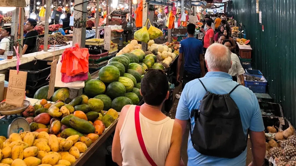 Voyageurs dans l’allée d’un marché couvert de Kuala Lumpur, entre les étals de pastèques et de mangues